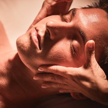Massage therapist gently massages sides of man's face as he relaxes with his eyes closed