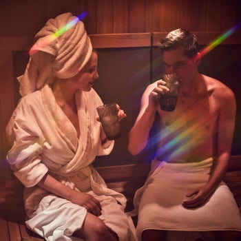 Couple sitting side-by-side in sauna and drinking iced cucumber water