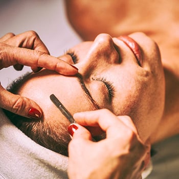 Aesthetician using a medical grade scalpel to remove fine hairs and dead skin from a woman's head during a dermaplaning facial.