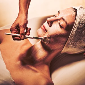 A woman relaxing on a table face-up while an esthetician brushes a mask on her face during a facial package.