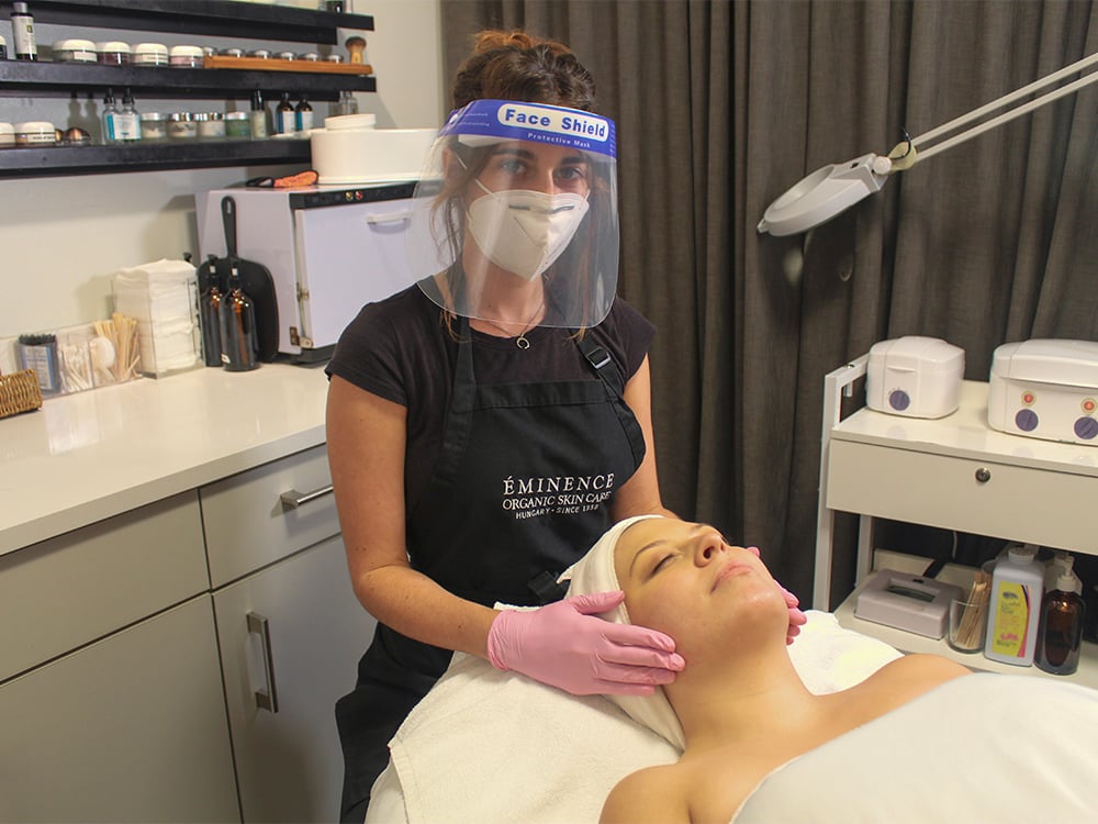 Aesthetician wearing a mask and face shield during a facial treatment with a client.