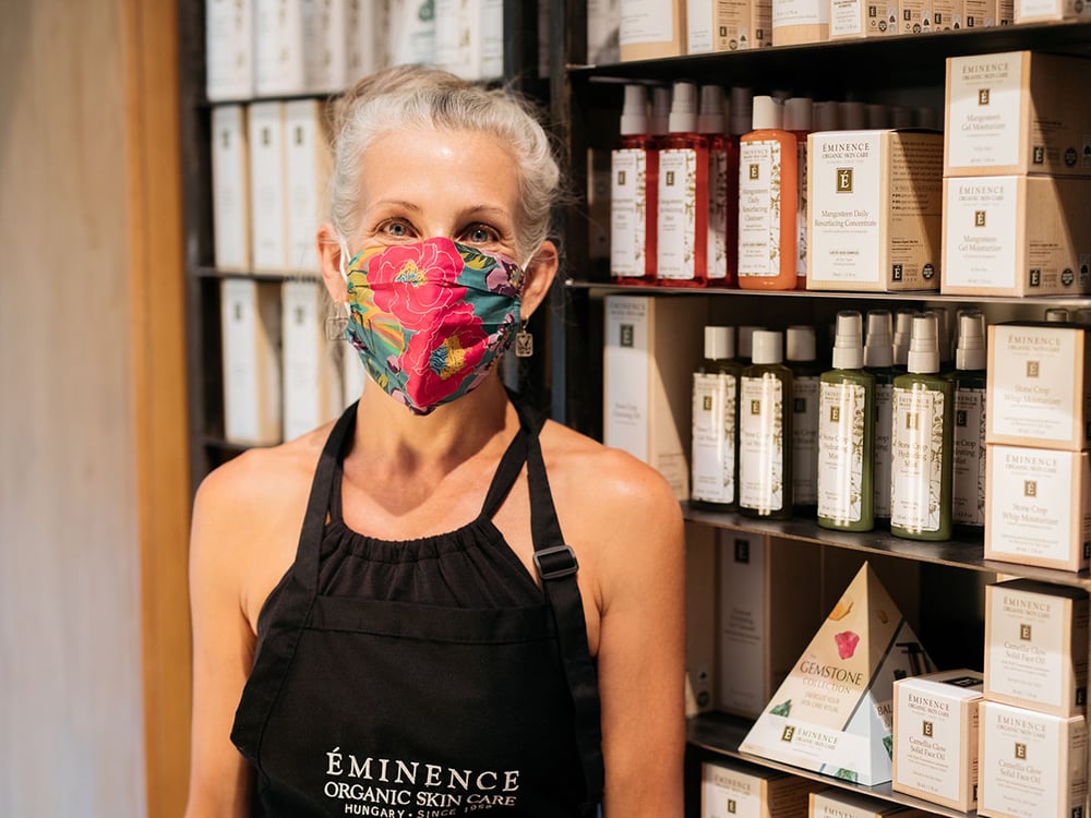 Aesthetician wearing K-95 mask standing in front of Eminence Organics products at Viva Day Spa