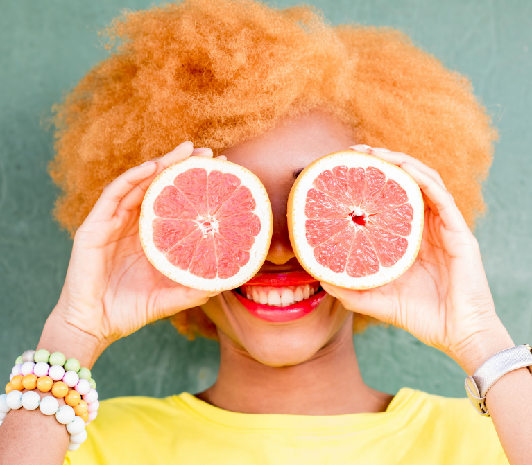 Close-up of smiling woman holding two grapefruit halves in front of her eyes.