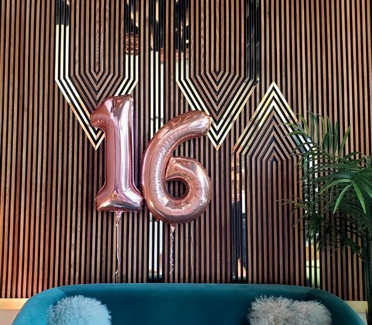 Large pink 16 balloons floating in front of Viva Day Spa's decorative wall to celebrate the spa's 16th anniversary in Austin, Texas.