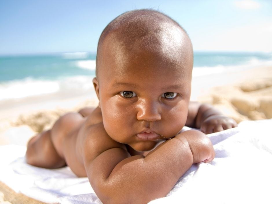 Baby lying on a towel in the sunshine looking into the camera, with the ocean in the background.