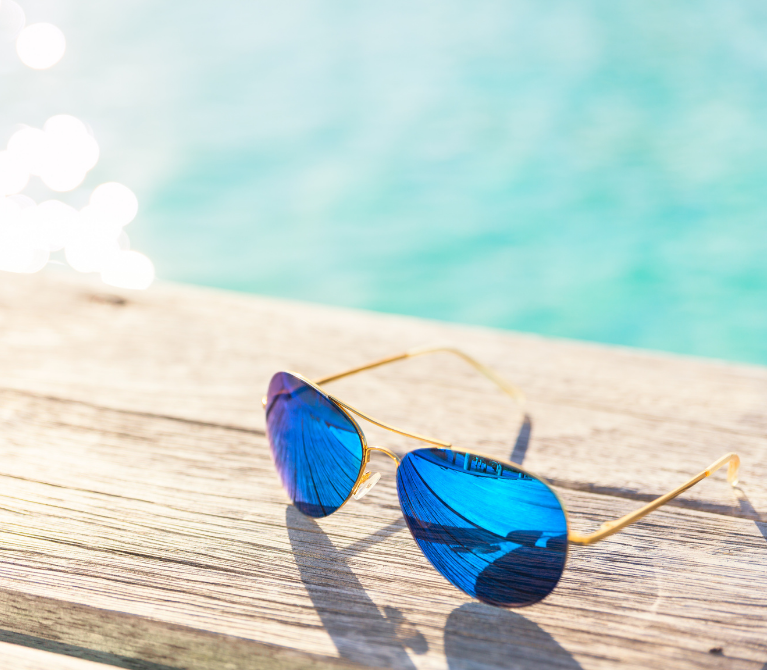 Pair of aviator sunglasses sitting on a wooden dock with sunshine reflecting off the water behind them.