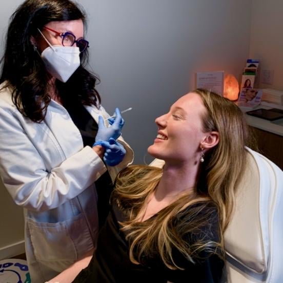 Aesthetic injector wearing a mask and gloves holds a syringe near the jawline of a smiling, reclined med spa client at Viva Day Spa + Med Spa.