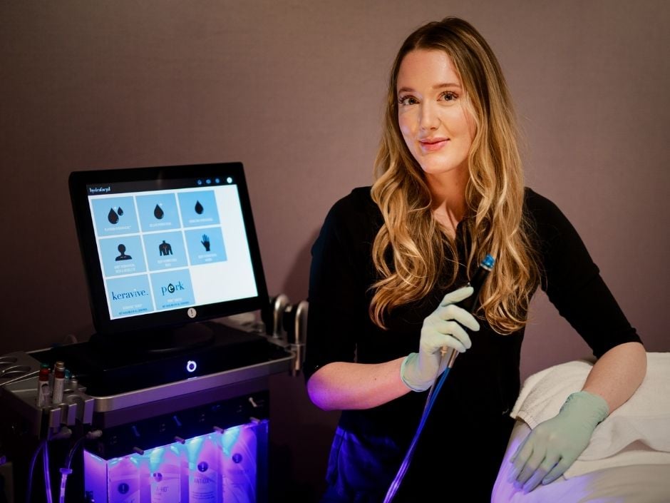 Viva Day Spa + Med Spa medical esthetician, Raya Kern, sitting next to a Hydrafacial system and looking into the cameral\.