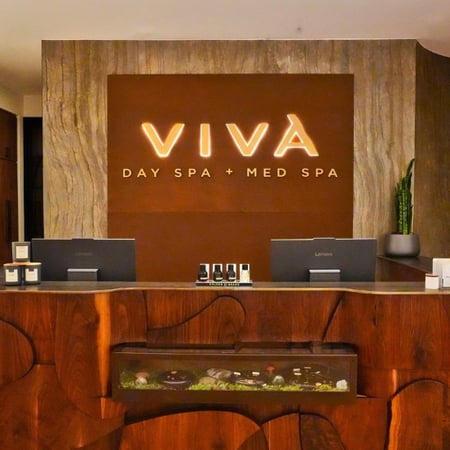 Warm wood reception desk with illuminated Viva Day Spa + Med Spa signage and display case at the Dallas, TX location